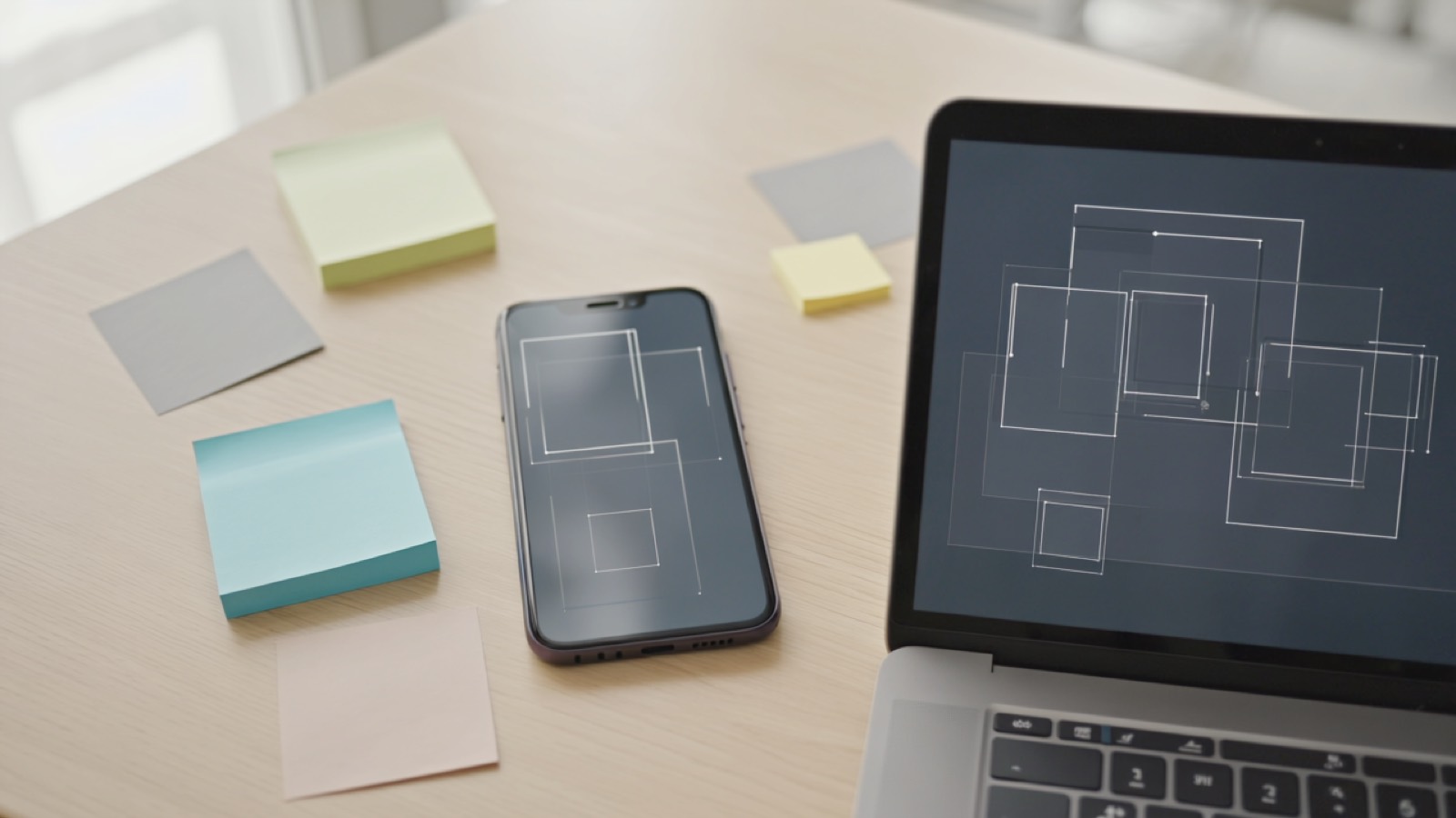 Smartphone und Laptop nebeneinander auf einem Schreibtisch, beide mit abstrakten Wireframe-Layouts und Notizzetteln